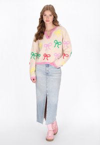 Strickpullover mit mehrfarbigen Schleifenmustern, V-Ausschnitt, rosa Besatz; kombiniert mit einem langen Jeansrock mit einem Schlitz vorne und rosa Sneakers.