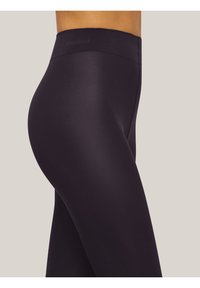 Primo piano di una persona che indossa leggings neri a vita alta e senza cuciture, su uno sfondo neutro uniforme.