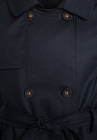 Manteau de marine avec un design croisé, muni de boutons marron, d'une ceinture nouée et d'un col classique en tissu lisse.