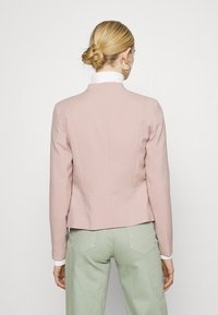 Femme aux cheveux blonds tressés portant des boucles d'oreilles anneaux dorés, un blazer rose, un col roulé blanc et un pantalon vert clair, vue de dos.