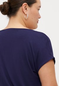 Camisa azul marino de manga corta, de tejido suave, con escote redondeado y costuras visibles, combinada con un pendiente de aro plateado.