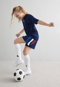 Camiseta y pantalones cortos deportivos de color marino con franjas rojas, azules y blancas; calcetines blancos y botas de fútbol; balón de fútbol en el suelo; fondo gris claro.