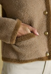 Bež herringbone-mustriga kardigan pehme tekstuuriga, pruunide ääriste ja kuldsete nööpidega; sisaldab küljetaskuid ja pehme viimistlusega.