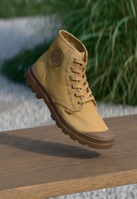 Botas altas de caña fabricadas en lona de color marrón, con emblema redondo, diseño con cordones y suela de goma marrón con un patrón de agarre texturizado.