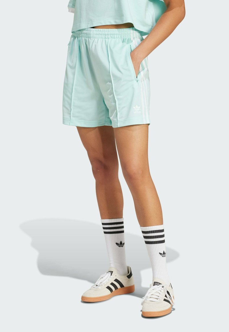 Pantalones cortos deportivos de un verde menta claro con una cintura elástica, bolsillos laterales y acentos de tres rayas blancas. Combinados con calcetines blancos a rayas y zapatillas.