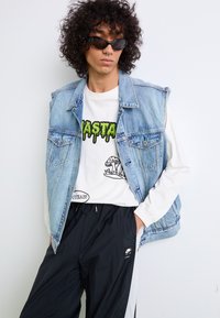 Gilet en denim bleu clair avec des emmanchures effilochées ; chemise à manches longues blanche avec un graphique vert ; pantalon de sport noir avec une bande latérale et un logo.
