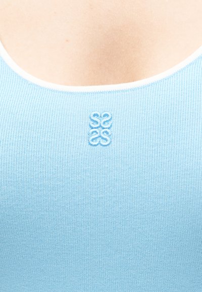 Κλείσιμο εικόνας ενός ατόμου που φοράει ένα ανοιχτό μπλε ριμπ πλεκτό τοπ με ένα μικρό ανοιχτό μπλε κεντημένο λογότυπο στο κέντρο κάτω από το λαιμόκοψη.