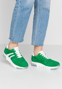 Sneaker in suede verde con dettagli e lacci bianchi, dotate di una suola imbottita e una finitura testurizzata. Indossate con jeans di denim risvoltati.