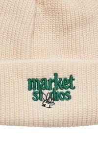Krémová čepice vyrobená z ribkované pleteniny. Obsahuje zelený vyšitý nápis "market studios" a grafiku ruky.
