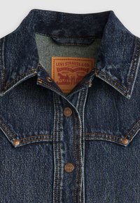 Tumedast sinisest denimist jakk, millel on kaks eesmist taskut, vasknõelad ja silt, millel on pruunis kirjas "Levi Strauss & Co."