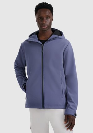 Mann trägt eine blaue Kapuzenjacke mit Reißverschluss, die Hände in den Taschen, steht vor einem einfarbigen hellen Hintergrund.