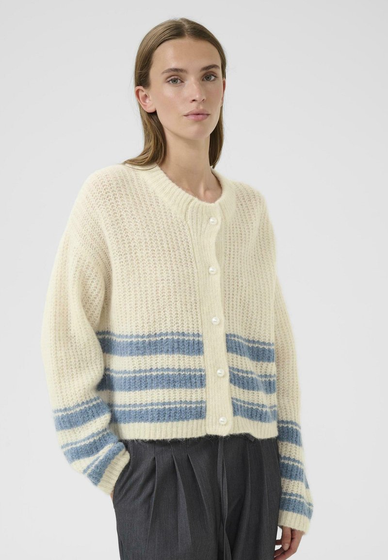 Eine Frau trägt eine gestrickte cremefarbene Cardigan mit blauen Horizontalstreifen und Perlenknöpfen, kombiniert mit dunklen, gefalteten Hosen, vor einem hellen Hintergrund.