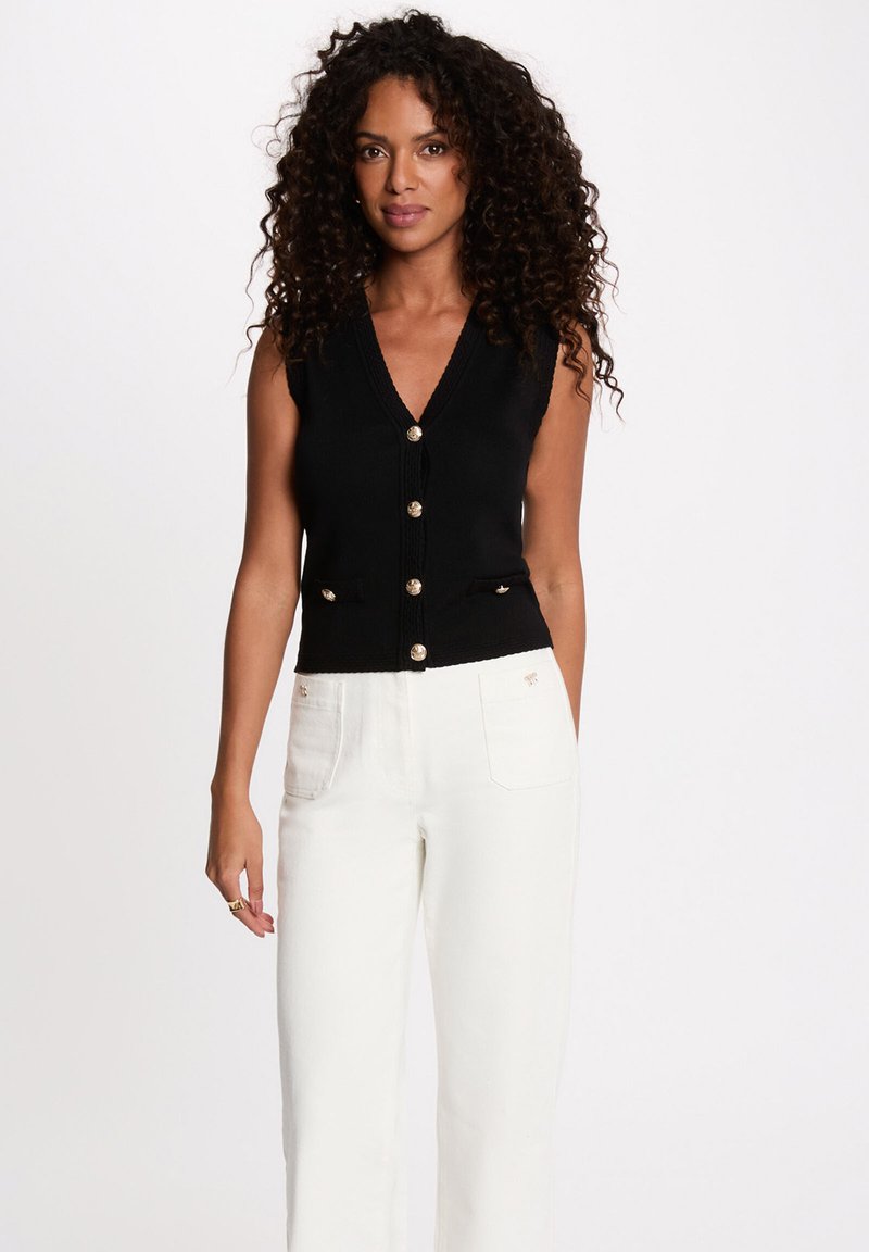 Woman with curly hair wearing sleeveless black buttoned vest and high-waisted white pants, standing against white background.