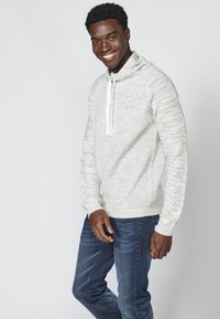 Hellgrauer gestrickter Pullover mit Kragen und weißem Zugband. Lange Ärmel mit gerippten Bündchen. Getragen mit dunkelblauen Jeans.