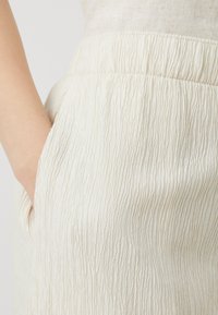 Pantalones de textura crema con ondas verticales, que cuentan con un bolsillo lateral y una cinturilla elástica, fabricados en un tejido ligero.
