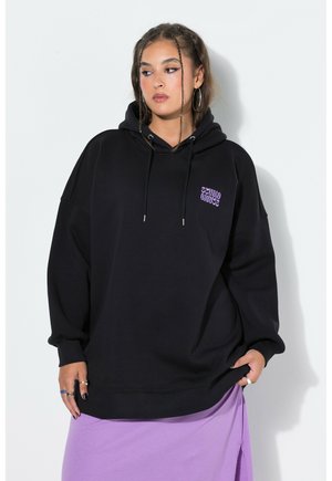 Femme avec des tresses portant un sweat à capuche noir avec un texte violet et une jupe lavande, debout sur un fond clair, regardant de côté.