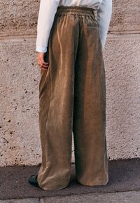 Pantaloni kaki largi din catifea cu un model texturat în dungi verticale și talie elastică. Purtate cu pantofi slip-on negri.