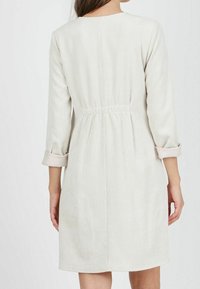 Nainen, jolla on yllään vaaleanbeige ribatty pitkähihaisia mekko, jonka hihansuut on käännetty ylös ja vyötärö on kiristetty, kuvattuna selkäpuolelta.