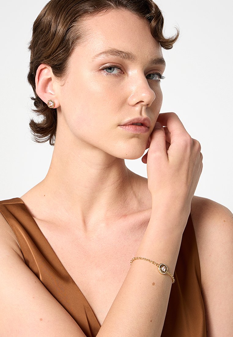 Pulsera de oro con una gema redonda, acompañada de pendientes de botón redondos. La modelo lleva un vestido de satén marrón, mostrando joyas delicadas contra un fondo neutro.