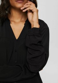 Blouse noire avec décolleté en V, fermeture à boutons, texture douce et manches bouffantes froncées avec détails aux poignets.