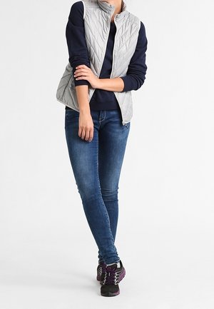 Graue gesteppte Weste mit hohem Kragen über einem marineblauen Sweatshirt, kombiniert mit blauen Jeans und Turnschuhen mit lila Akzenten.