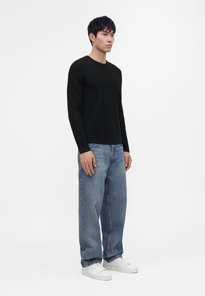 Homme debout portant une chemise noire à manches longues, un jean bleu large et des baskets blanches sur un fond blanc uni.
