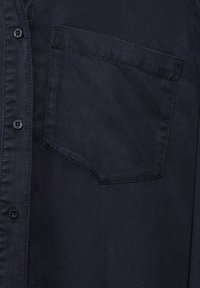 Tmavě námořnická košile na knoflíky s texturovanou tkaninou, jednou náprsní kapsou a černými plastovými knoflíky podél předního zapínání.