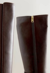 Pereche de cizme înalte din piele maro închis, cu cusături vizibile și un fermoar auriu pe o cizmă, așezate pe un fundal deschis.