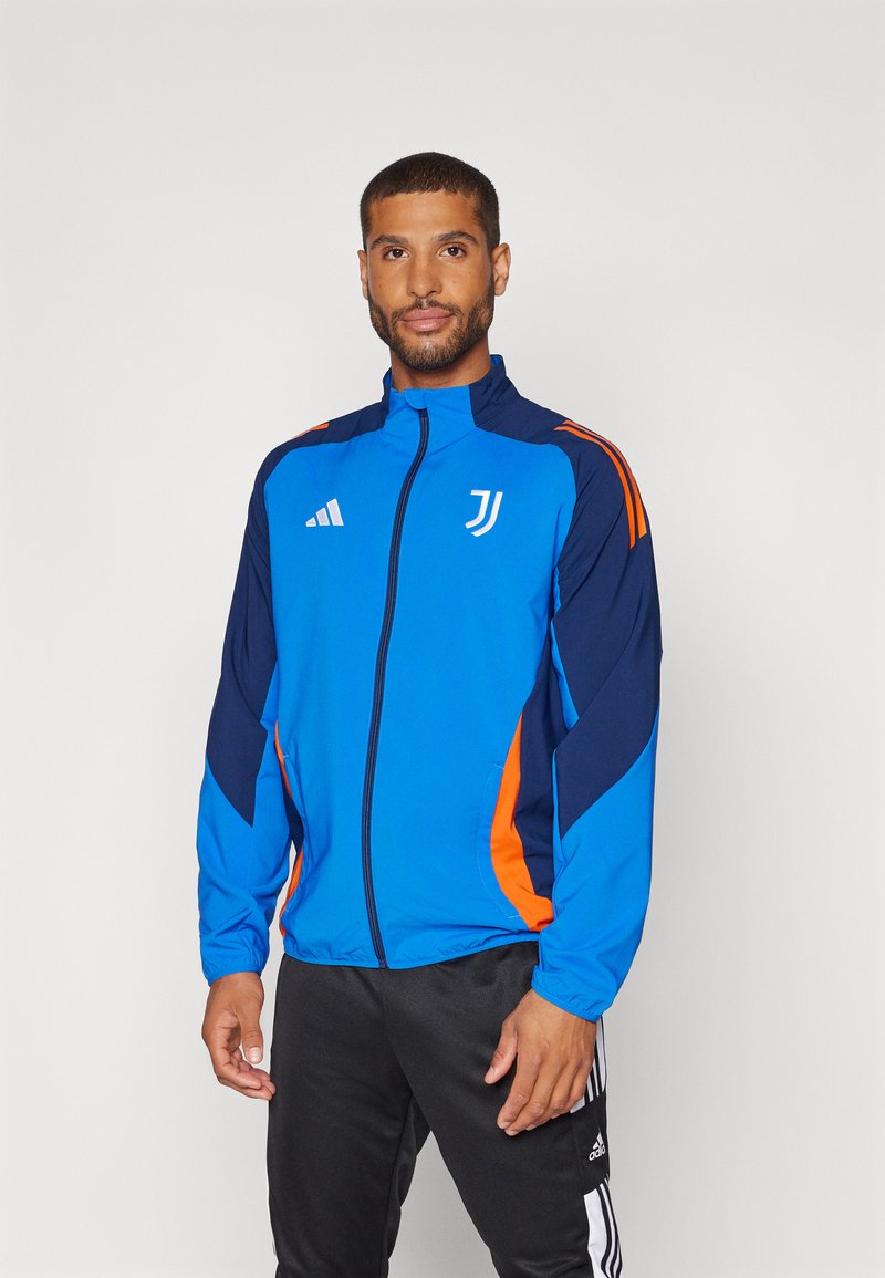 Chaqueta azul con cierre de cremallera y detalles en navy y naranja, con un alto collar, dos bolsillos laterales y el logo de la Juventus en el pecho.