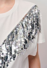 Witte T-shirt met een diagonale franjerand van zilveren pailletten, wat een textuurcontrast langs één kant creëert. Korte mouwen, ronde halslijn.