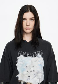Camiseta negra de gran tamaño con un estampado gráfico de dos lobos blancos entre nubes. El texto en la parte superior dice "BIMBAYLOLA". Tela de algodón con un corte holgado.