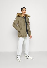 Parka verde oliva con cappuccio in pelliccia sintetica, cordini e molteplici tasche. Indossata sopra una maglietta nera, abbinata a jeans bianchi e scarpe sportive in bianco e nero.