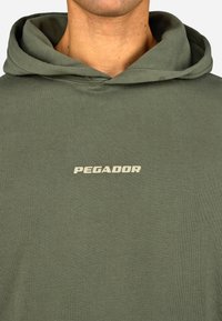 Sudadera verde con capucha hecha de una tela suave, que presenta una textura acanalada y un logotipo en crema en la parte delantera que dice "PEGADOR."