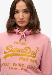 Roze hoodie met een trekkoordkap en gouden glitters die "Vintage SuperDry" uitstralen. Zacht materiaal, casual pasvorm en gestikte accenten.