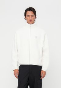 White fleece jacket with a high collar and front zipper. Features visible stitching, a logo on the chest, and side pockets.
