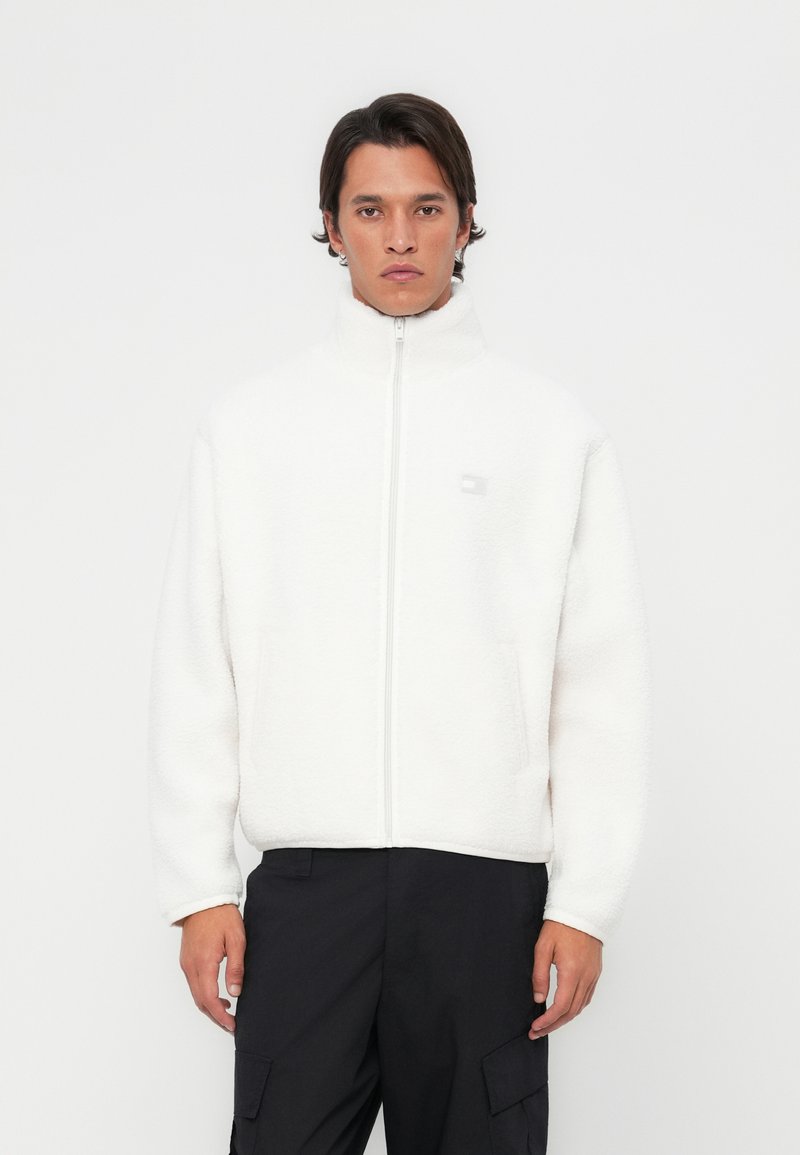 White fleece jacket with a high collar and front zipper. Features visible stitching, a logo on the chest, and side pockets.