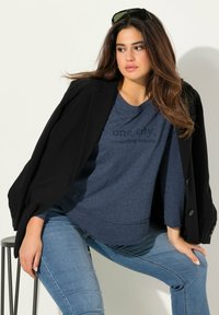 Schwarzer Blazer über einem blauen Sweatshirt mit geprägtem Text, kombiniert mit hellblauen Jeans. Model sitzt auf einem Hocker vor einem neutralen Hintergrund.