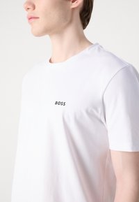 Tricou alb din bumbac, cu mâneci scurte, o gâtă rotundă și un mic logo negru "BOSS" pe piept. Textură netedă și ușoară.