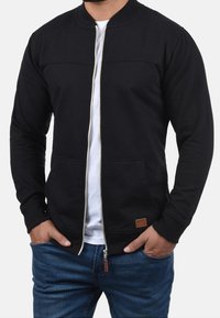 Musta vetoketjullinen takki, jossa pyöreä kaula-aukko, teksturoidulla yläosalla ja kahdella sivutaskulla. Päällä valkoinen t-paita. Farkut näkyvissä.