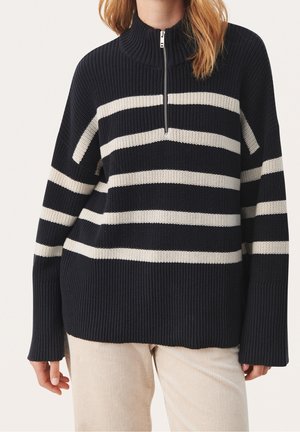 Kobieta w czarnym swetrze dzianym z białymi poziomymi paskami i wysokim kołnierzem na półzamek, w połączeniu z jasnobeżowymi spodniami sztruksowymi.