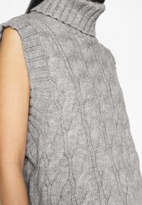 Suéter gris de punto trenzado con un alto cuello de tortuga acanalado. El diseño sin mangas presenta patrones texturizados a lo largo de la tela. Apariencia suave y acogedora.