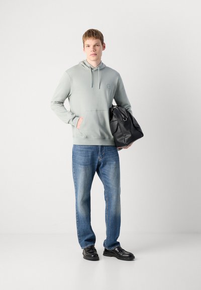 Jeune homme portant un sweat à capuche gris clair, un jean bleu et des chaussures noires, tenant un grand sac en cuir noir contre un fond blanc uni.