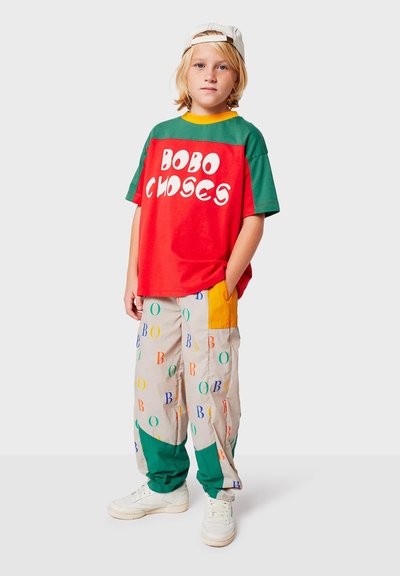Rode en groene shirt met korte mouwen met witte tekst "BOBO CHOSES." Beigetinten broek met multikleurige letters en patches. Witte sneakers en pet.