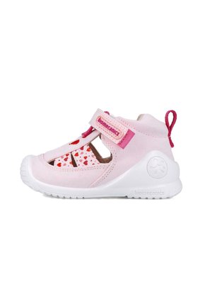 Sandal de tela rosa con patrones de corazones recortados, suela de goma blanca, correa de velcro ajustable y una pestaña en el talón para facilitar su uso.