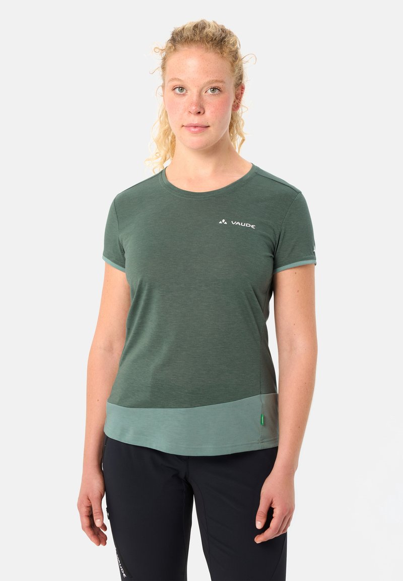Vrouw met krullend blond haar draagt een groen sportshirt met korte mouwen en het logo van "Vaude", en zwarte broek, staand tegen een effen achtergrond.