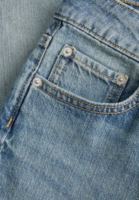 Jeans de mezclilla con un lavado azul claro, que presentan costuras visibles, un pequeño bolsillo frontal y acentos en hardware plateado. Tela texturizada.