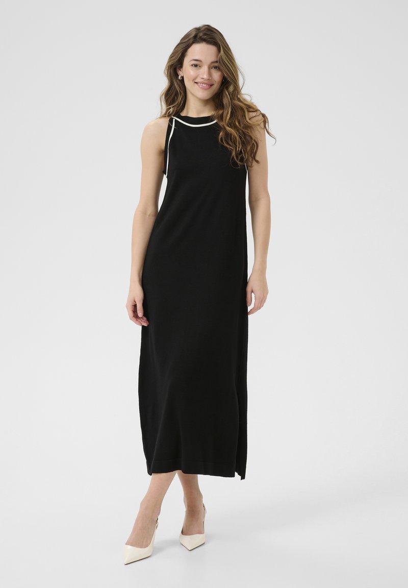 Robe maxi noire sans manches avec bord blanc au niveau du col ; coupe droite ; tissu doux ; fente latérale haute ; portée avec des talons aiguilles crème.