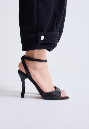 Pie femenino con sandalia negra de tacón alto con correa en el tobillo, combinado con pantalones negros cortos con puño abotonado.