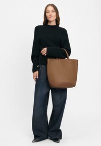 Bruine leren tote bag met een gladde textuur en gestructureerde vorm. Voorzien van korte handvatten en minimale branding. Draagt met een zwarte trui en jeans.