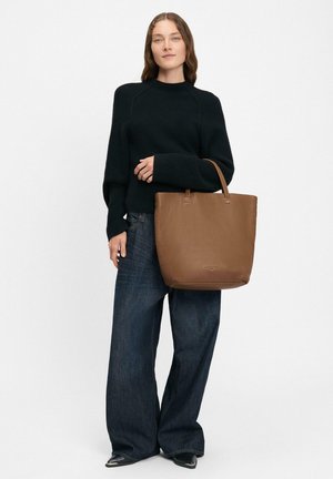 Borsa tote in pelle marrone con una trama liscia e una forma strutturata. Caratterizzata da manici corti e minimo branding. Indossata con un maglione nero e jeans.